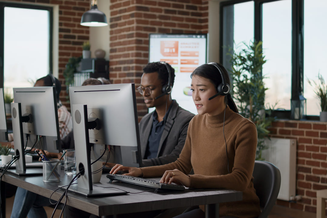 Funcionários de um call center trabalham em um ambiente moderno e bem iluminado. Em primeiro plano, uma mulher usando um suéter marrom e um headset conversa atenta enquanto digita no teclado. Ao fundo, outros atendentes, incluindo um homem de óculos e terno, estão concentrados em seus computadores. A sala tem uma estética industrial, com paredes de tijolos aparentes, plantas e janelas amplas.