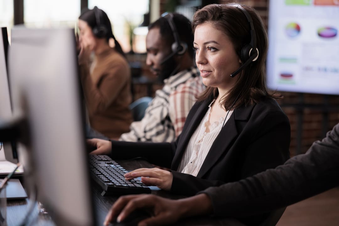 Funcionários de um call center estão trabalhando em uma sala moderna e bem iluminada. Uma mulher em destaque, vestindo um blazer preto e com um headset, está concentrada em seu monitor enquanto digita. Outros colegas aparecem em segundo plano, usando headsets e trabalhando em computadores, em um ambiente profissional com gráficos visíveis ao fundo.