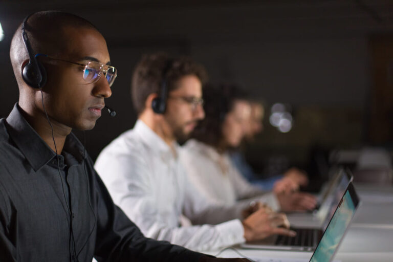 A imagem mostra uma linha de pessoas trabalhando em computadores, todas usando fones de ouvido com microfones, sugerindo que estão em um call center ou central de atendimento. A pessoa em destaque está à esquerda da imagem, vestindo uma camisa preta e óculos, concentrada no monitor de seu laptop. Ao fundo, outras pessoas, também com fones de ouvido e em frente a computadores, estão um pouco desfocadas, mas também parecem estar focadas em suas tarefas. A iluminação é suave, destacando um ambiente de trabalho em local fechado e controlado.