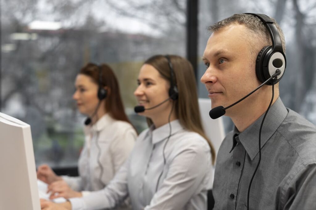 A imagem mostra três profissionais em um call center, usando fones de ouvido com microfone, sentados lado a lado e aparentemente concentrados em seus trabalhos. Eles estão olhando para as telas de seus computadores, com expressões focadas e amigáveis. Ao fundo, uma janela ampla permite a entrada de luz natural, revelando uma vista externa levemente desfocada de árvores e prédios. Essa cena sugere um ambiente de trabalho colaborativo e orientado ao atendimento ao cliente.