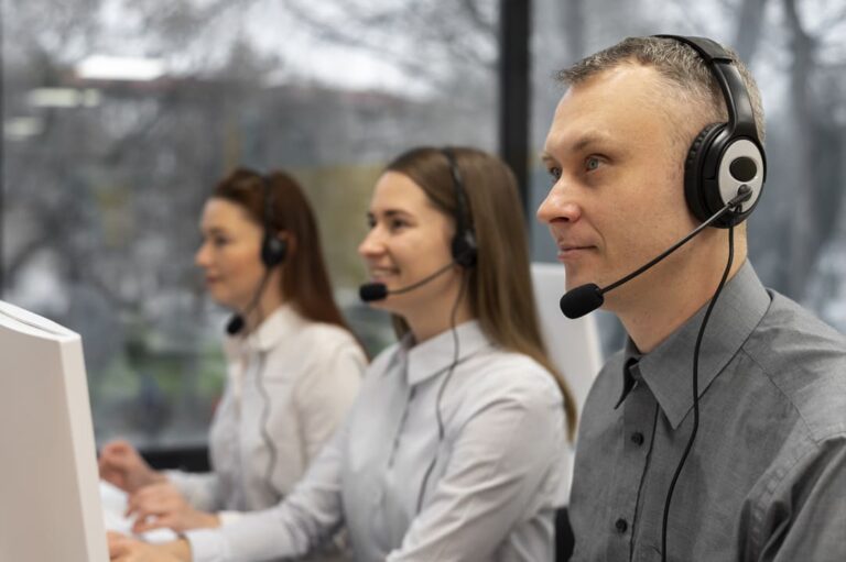 A imagem mostra três profissionais em um call center, usando fones de ouvido com microfone, sentados lado a lado e aparentemente concentrados em seus trabalhos. Eles estão olhando para as telas de seus computadores, com expressões focadas e amigáveis. Ao fundo, uma janela ampla permite a entrada de luz natural, revelando uma vista externa levemente desfocada de árvores e prédios. Essa cena sugere um ambiente de trabalho colaborativo e orientado ao atendimento ao cliente.