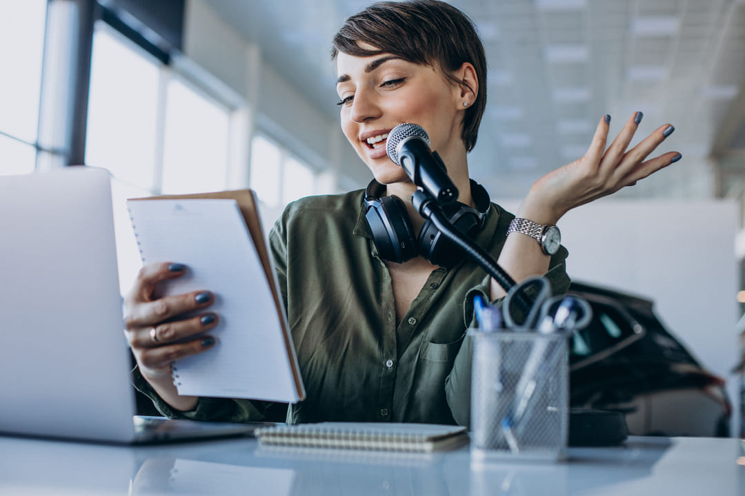 Uma mulher jovem, sentada em uma mesa, grava um podcast ou realiza uma locução. Ela segura um bloco de notas, fala no microfone e usa um par de fones de ouvido pendurados no pescoço.